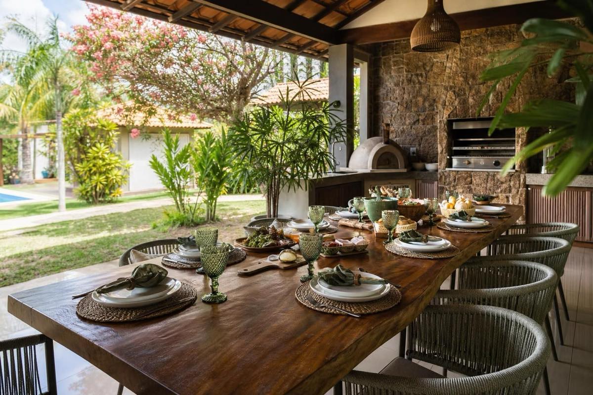 Mesa comprida posta em área gourmet coberta com churrasqueira, vista para jardim tropical ensolarado.