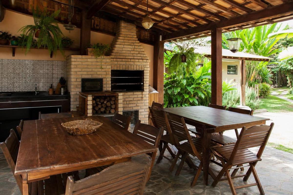 Área gourmet coberta com churrasqueira de alvenaria e mesas de madeira em ambiente aberto com jardim tropical.