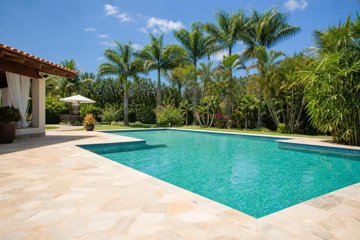 Piscina ampla e vazia com deck de pedra sob sol da manhã, coqueiros ao fundo e jardim tropical